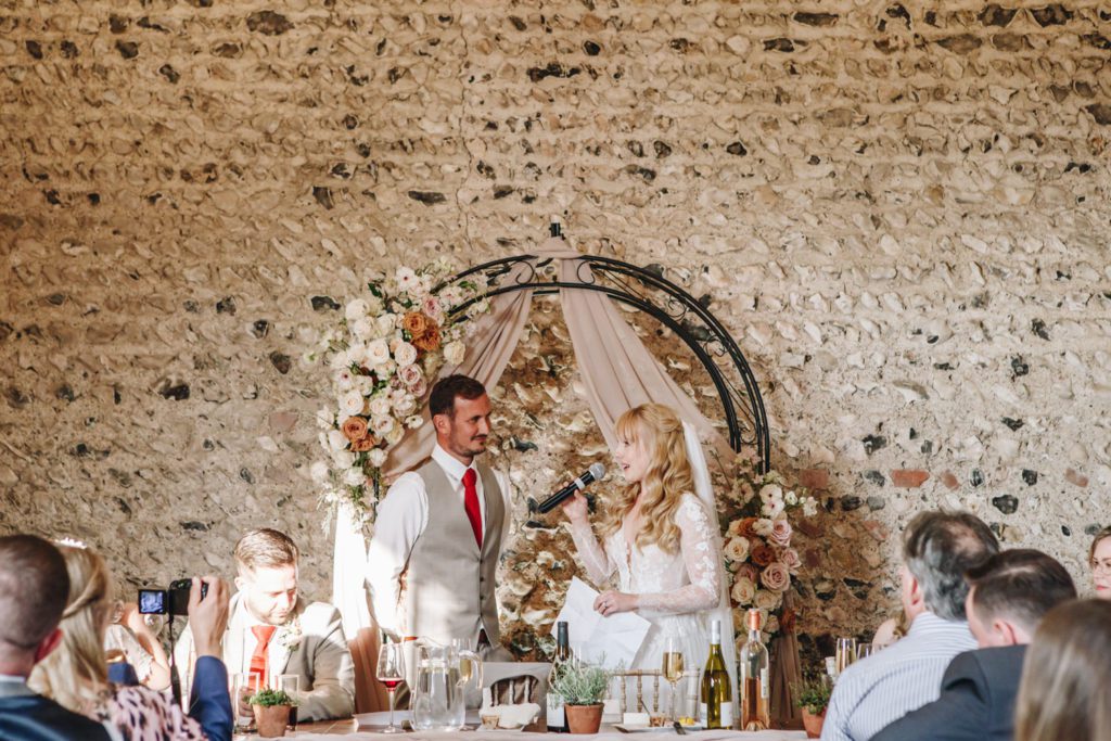 A couple celebrating their wedding at Cissbury Barns standing in front of a romantic wedding flower archway during their speeches